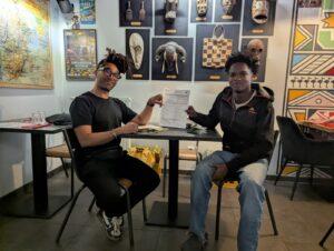 Dans le restaurant Jabiru Café, en arrière plan, les murs sont couverts d'affiches et de décorations rappelant les cultures d'Afrique. Au premier plan, tournés vers l'objectif, le propriétaire du café et Muhammed alim, jeune accueilli au centre, sont assis face à face. Ils affichent tous les deux un grand sourire et tiennent chacun le contrat en le montrant à l'objectif.