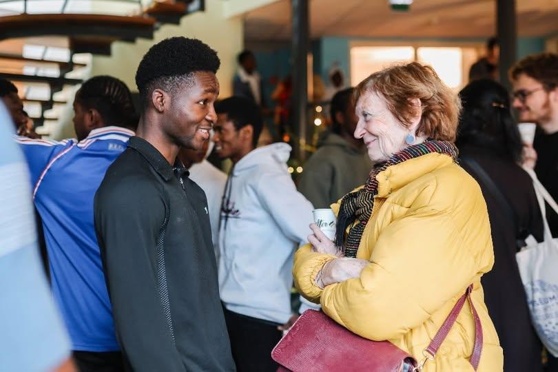 Dans l'entrée du Centre Bernanos, nous voyons un groupe de personnes, mélangeant jeunes accueillis au centre, bénévoles, et visiteurs extérieurs, qui se rencontrent et discutent. Au premier plan, un jeune homme et une femme plus agée s'échangent un sourire, ils semblent discuter et passer un bon moment.